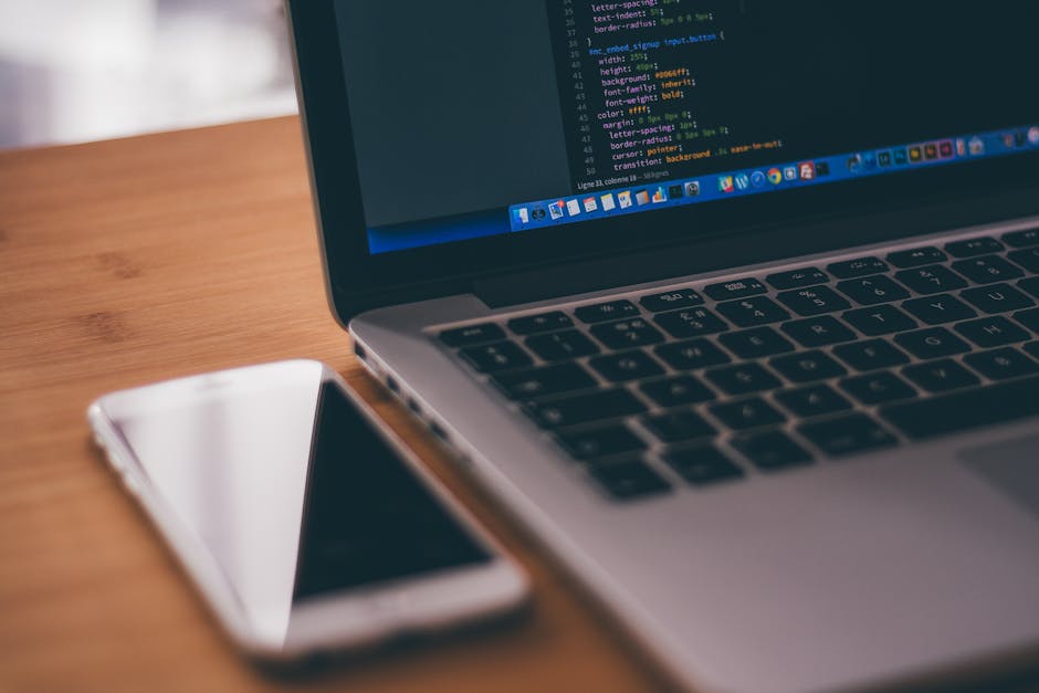 A close-up view of a laptop on a wooden desk displaying lines of computer code on its screen, with a white smartphone placed to the left of the laptop. The laptop's keyboard and touchpad are visible, 