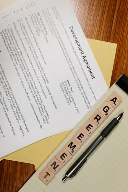 A close-up view of a development agreement document laid out on a wooden surface, partially covered by a yellow folder. Next to the document is a black and silver pen, and a notepad with blank lines.